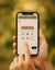 Eine Hand hält ein iPhone mit geöffneter Upload-Seite von magnet-momente.de, auf der ein Button zum Bilderauswählen sowie mehrere Plattform-Icons zu sehen sind. Der Hintergrund ist weich und warm verschwommen